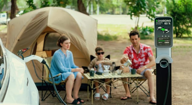 Outdoor adventure and family vacation camping in nature travel by eco friendly car for sustainable future. Lovely sit on trunk, charging EV car with EV charging station in campsite. Panorama Perpetual