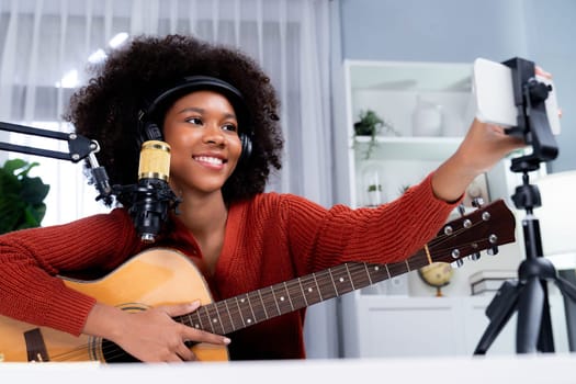 Host channel of beautiful African woman influencer setting smartphone, sing with play guitar in broadcast. Time slot of music blogger on live social media online. Concept of audio creator. Tastemaker.