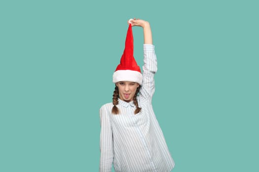 Portrait of happy funny teenager girl with braids wearing striped shirt, looking at camera with tongue out, pulling up her Santa Claus hat. Indoor studio shot isolated on green background.