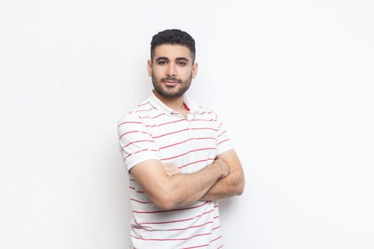 Portrait of delighted positive confident attractive bearded man wearing striped t-shirt standing with crossed hands, looking at camera. Indoor studio shot isolated on gray background.