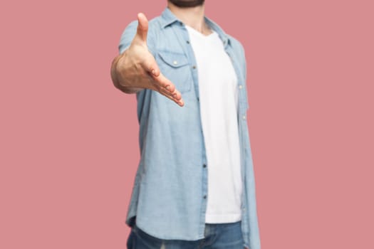 Unknown man in blue casual style shirt standing giving hand for handshake as sign of greeting, saying hello to person he meeting. Indoor studio shot isolated on pink background.