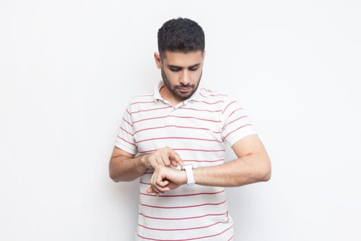 Portrait of concentrated attractive handsome bearded man wearing striped t-shirt standing using his smart watch, uses application. Indoor studio shot isolated on gray background.