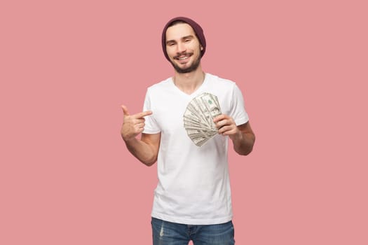 Portrait of satisfied delighted bearded man in white T-shirt and beany hat standing pointing at dollar banknotes, looking smiling at camera. Indoor studio shot isolated on pink background.