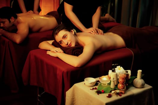 Caucasian couple customer enjoying relaxing anti-stress spa massage and pampering with beauty skin recreation leisure in warm candle lighting ambient salon spa at luxury resort or hotel. Quiescent