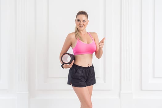 Portrait of sporty young beautiful woman with perfect body wearing pink top and black shorts standing, holding foam roll and showing thumb up, looking at camera. Indoor studio shot.