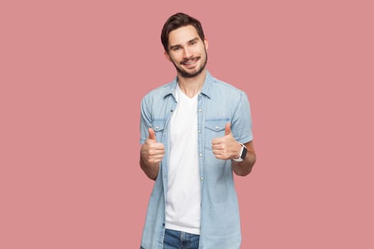 Man in blue shirt gesturing thumbs-up, telling you that you are doing a good job, looking and smiling at the camera with cheerful expression. Indoor studio shot isolated on pink background.