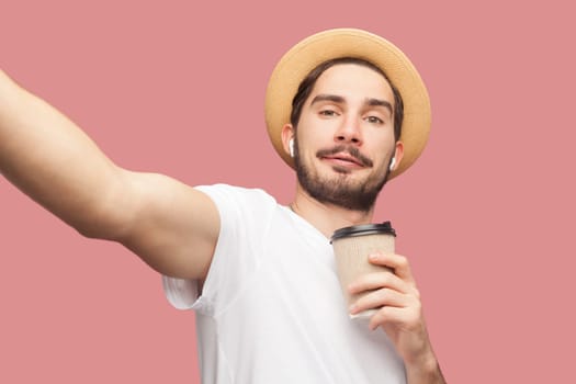 Portrait of smiling positive attractive bearded man blogger in white T-shirt and hat standing with coffee and making pointy of view photo. Indoor studio shot isolated on pink background.