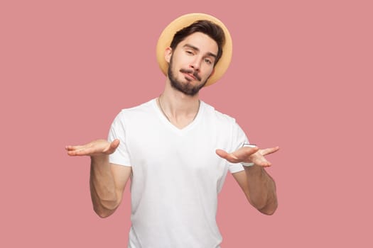 Portrait of confident handsome attractive bearded man in white T-shirt and hat standing looking at camera, showing rock cool gesture, looks playful. Indoor studio shot isolated on pink background.