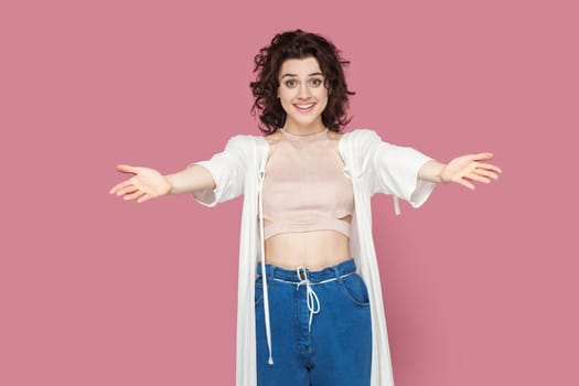 Welcome. Portrait of extremely happy pretty woman with curly hair wearing casual style outfit standing with outstretched hands, inviting. Indoor studio shot isolated on pink background.