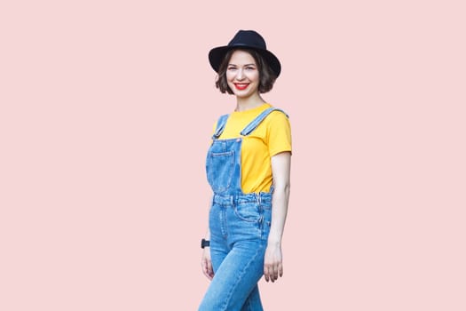 Portrait of cheerful positive hipster woman in blue denim overalls, yellow T-shirt and black hat, looking at camera with toothy smile. Indoor studio shot isolated on light pink background.