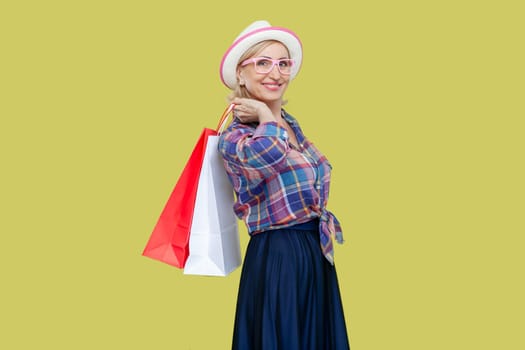 Side view portrait of joyful cheerful mature woman wearing checkered shirt, hat and eyeglasses holding shopping bag, enjoying. Indoor studio shot isolated on yellow background.