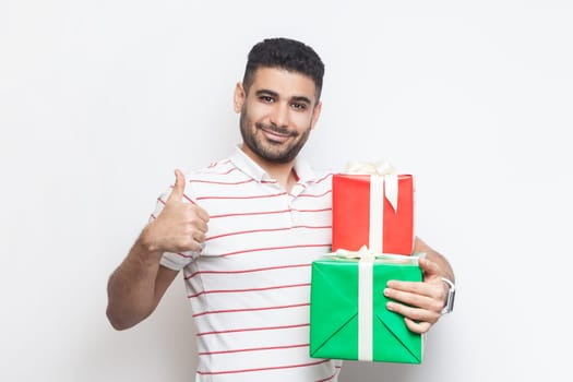 Portrait of attractive cheerful joyful bearded man wearing striped t-shirt standing looking at camera, holding two gift boxes, showing like gesture. Indoor studio shot isolated on gray background.
