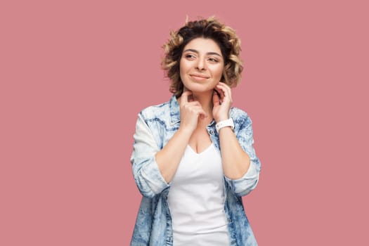 Portrait of smiling beautiful woman with curly hairstyle wearing blue shirt standing looking away, smiling, dreaming about something pleasant. Indoor studio shot isolated on pink background.