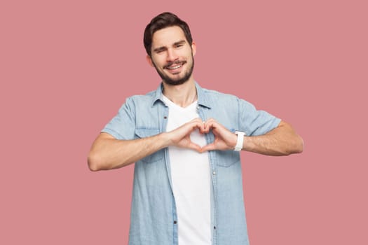 Portrait of bearded man in blue casual style shirt standing showing heart gesture over chest, being passionate, express love to close person. Indoor studio shot isolated on pink background.