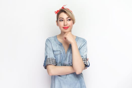 Portrait of positive smiling optimistic blonde woman wearing blue denim shirt and red headband standing looking away, dreaming. Indoor studio shot isolated on gray background.