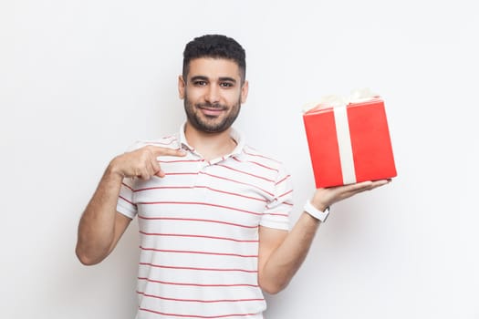Portrait of smiling positive optimistic bearded man wearing striped t-shirt standing holding red wrapped present box, pointing at gift. Indoor studio shot isolated on gray background.