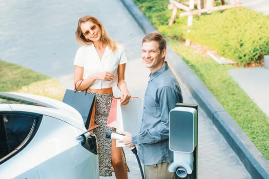 Young couple travel with EV electric car charging in green sustainable city outdoor garden in summer shows urban sustainability lifestyle by green clean rechargeable energy of electric vehicle innards