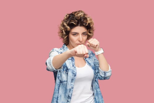 Portrait of angry aggressive woman with curly hairstyle wearing blue shirt standing with clenched fist, boxing, knocks somebody. Indoor studio shot isolated on pink background.