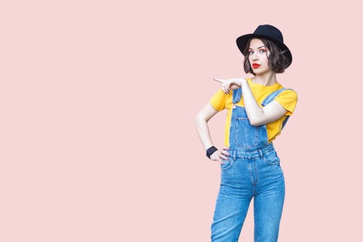 Portrait of surprised amazed hipster woman in blue denim overalls, yellow T-shirt and black hat, pointing at copy space for advertisement. Indoor studio shot isolated on light pink background.