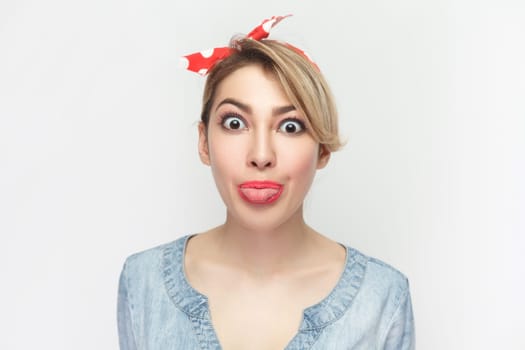 Portrait of funny crazy foolish blonde woman wearing blue denim shirt and red headband standing sticking tongue out, looking at camera. Indoor studio shot isolated on gray background.