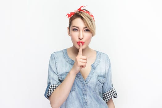 Portrait of serious concentrated blonde woman wearing blue denim shirt and red headband standing with finger near lips, keeps silence. Indoor studio shot isolated on gray background.