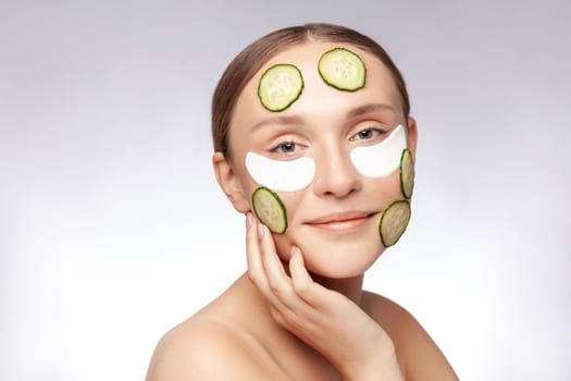Closeup portrait of smiling attractive beautiful woman making skin care procedures, applying under eyes patches and cucumbers on face. Indoor studio shot isolated over gray background.