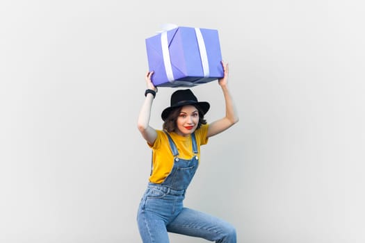 Portrait of funny joyful hipster woman in blue denim overalls, yellow T-shirt and black hat, holding big present box above her head, looking at camera. Indoor studio shot isolated on gray background.
