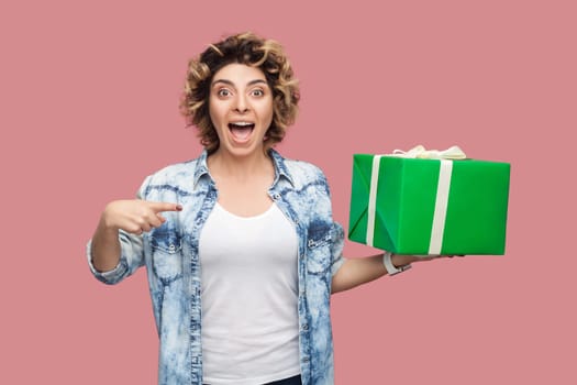 Portrait of excited happy beautiful woman with curly hairstyle wearing blue shirt pointing at green present box, being surprised, celebrating birthday. Indoor studio shot isolated on pink background.