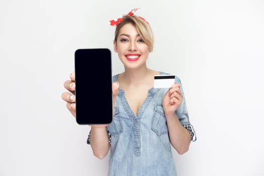 Portrait of joyful smiling blonde woman wearing blue denim shirt and red headband standing showing phone with empty display and credit card. Indoor studio shot isolated on gray background.