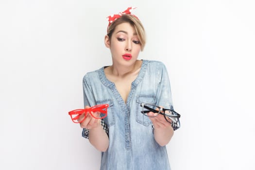 Portrait of confused blonde woman wearing blue denim shirt and red headband standing holding red and black glasses, can't choose better spectacles. Indoor studio shot isolated on gray background.