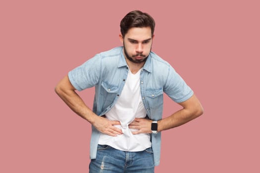 Portrait of bearded man in casual style shirt standing feels stomachache, suffers from indigestion and abdomainal cramps after eating spoiled product. Indoor studio shot isolated on pink background.
