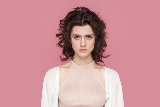 Portrait of woman with curly hairstyle wearing casual style outfit staring at camera with unhappy or regretful look, expressing sorrow and sadness. Indoor studio shot isolated on pink background.