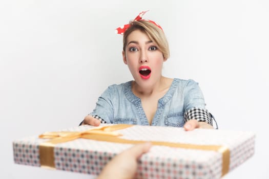 Portrait of astonished shocked blonde woman wearing blue denim shirt and red headband standing receives present box, being surprised. Indoor studio shot isolated on gray background.