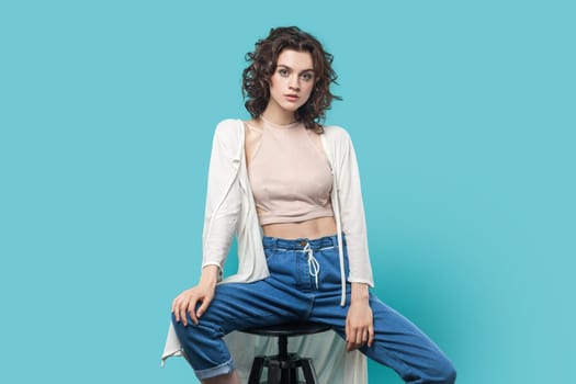 Portrait of self-confident beautiful brunette woman sitting on chair and posing like fashion model and looking at camera with serious face. Indoor studio shot isolated on blue background.