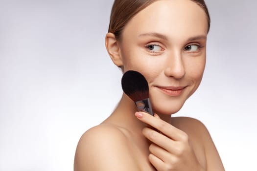 Closeup portrait of smiling adorable beautiful woman holding powder brush looking away, beauty procedure makeup. Indoor studio shot isolated over gray background.