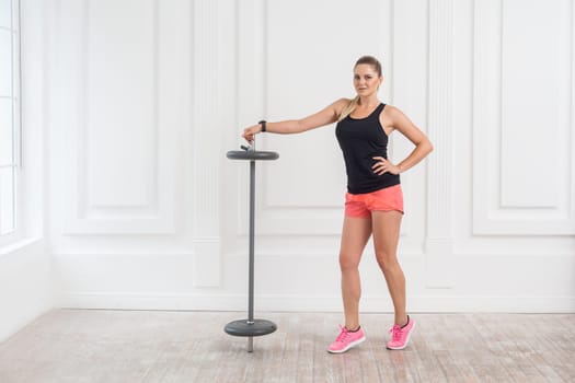 Full length portrait of athletic beautiful woman in pink top and black shorts standing, holding barbell and looking at camera with confident expression on white wall background. Indoor studio shot.