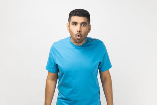 Portrait of amazed shocked surprised unshaven man wearing blue T- shirt standing looking at camera with big eyes, sees something astonished. Indoor studio shot isolated on gray background.