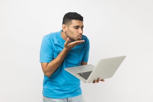 Portrait of romantic pleased unshaven man wearing blue T- shirt standing holding laptop, having video call, sending air kiss to his girlfriend. Indoor studio shot isolated on gray background.