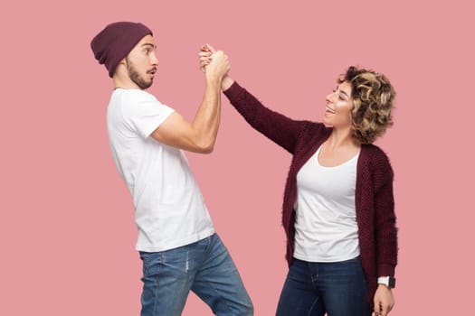 Portrait of smiling positive woman and surprised shocked man standing together and holding hands, looking at each other with different emotions. Indoor studio shot isolated on pink background.