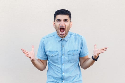 Portrait of aggressive crazy shocked handsome man wearing denim shirt standing with raised arms and screaming loud with hate and anger. Indoor studio shot isolated on gray background.