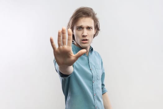 Portrait of young man making stop gesture showing palm of hand, conflict prohibition warning about danger, stop bullying, wearing blue shirt. Indoor studio shot isolated on gray background.