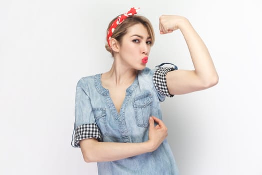 Portrait of strong powerful blonde woman wearing blue denim shirt and red headband standing raised her arm, showing biceps, keeps lips pout. Indoor studio shot isolated on gray background.