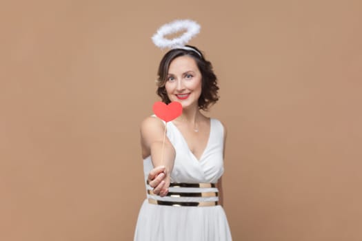 Portrait of angelic woman with wavy hair and nimb over head, giving little red hearts, congratulating with Valentines Day, wearing white dress. Indoor studio shot isolated on light brown background.