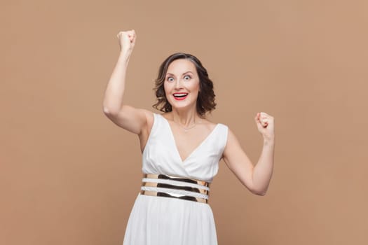 Portrait of extremely happy middle aged woman with wavy hair clenching fists and yelling happily, celebrating her triumph, wearing white dress. Indoor studio shot isolated on light brown background.