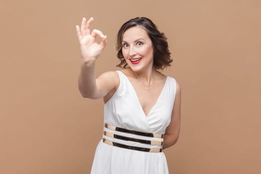 Everything is perfect. Portrait of happy positive woman with wavy hair showing OK gesture with hand, having good mood, wearing white dress. Indoor studio shot isolated on light brown background.