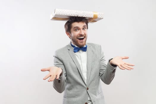 Portrait of joyful bearded man standing with present box on head, expressing happiness, saying take your gift, wearing grey suit and blue bow tie. Indoor studio shot isolated on gray background.