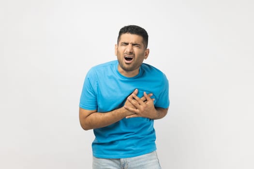 Portrait of stressed sick unshaven man wearing blue T- shirt standing with hands on chest, having heart attack, screaming from pain. Indoor studio shot isolated on gray background.