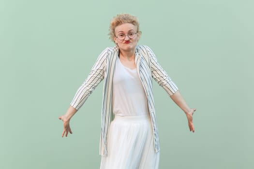 Portrait of confused puzzled young adult blonde woman wearing striped shirt and skirt, shrugging shoulders, doesn't know answer and what to do. Indoor studio shot isolated on light green background.