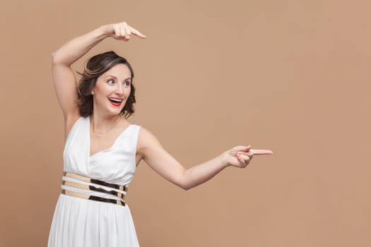 Portrait of amazed excited happy middle aged woman with wavy hair pointing aside at copy space for promotional text, wearing white dress. Indoor studio shot isolated on light brown background.