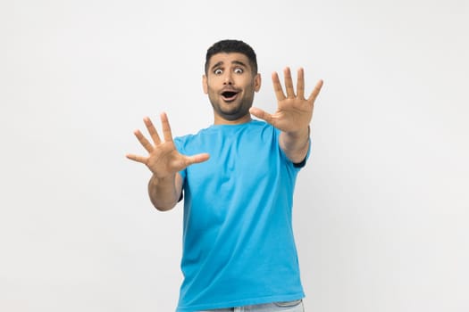 Portrait of scared shocked frighten unshaven man wearing blue T- shirt standing with outstretched hands to camera, looking with big eyes, being afraid. Indoor studio shot isolated on gray background.